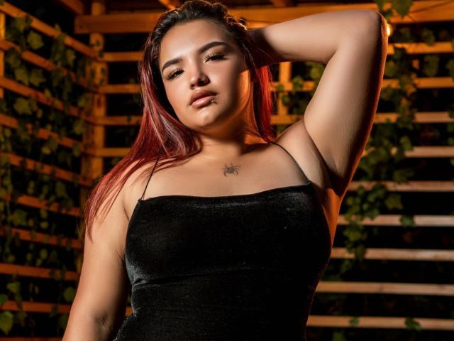 LilithMoonryn with red hair, black velvet dress, raising arm, showing armpit, small spider tattoo on chest, sexy, confident, soft lighting, warm colors, slight smile, dark background.
