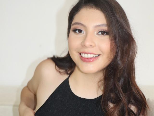 VioletaRoses, with brown wavy hair, smiling at the camera, wearing a black sparkly sleeveless top, showing her white teeth and pink lips.