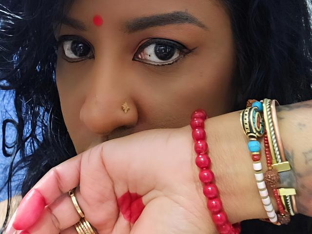 Nahirr's face with dark curly hair, brown skin, red dot on forehead, nose ring, and blue eyes, holding hand with red beads and multiple bracelets, pink nails.