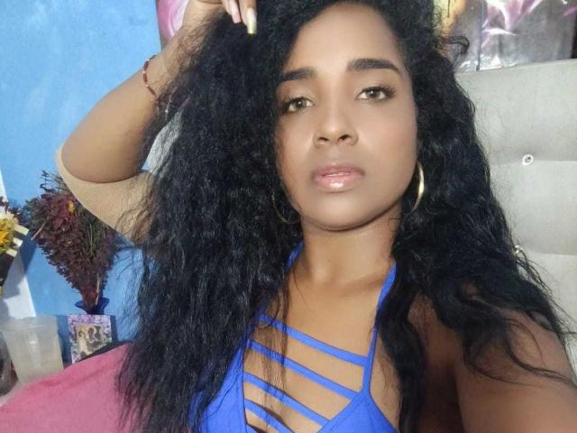 Selenabrowny with dark curly hair, wearing a blue strappy top, gold hoop earrings, and glossy lips, looking slightly to the side with a relaxed expression.