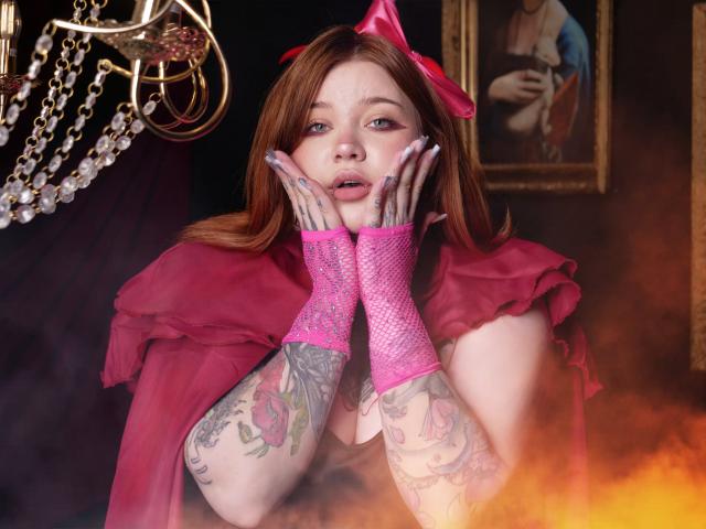 YoureGift, a fair-skinned woman with red hair, wears a pink bow and fishnet gloves. Her tattooed arms rest on her cheeks, hands slightly touching her face. She has pink lipstick and a surprised expression. She's dressed in a pink, ruffled top. A framed painting and a chandelier with chains are in the background. The lower right has a smoky, orange glow. Her tattoos include roses and floral designs. The image is sharp with vibrant colors.