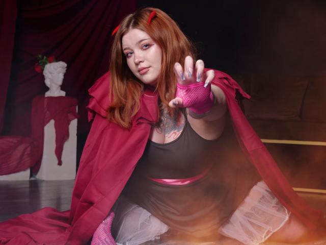 YoureGift with red hair, pink gloves, and a red cape, reaching out, showing her tattooed chest, wearing a black top and white skirt, in a sexy cosplay.