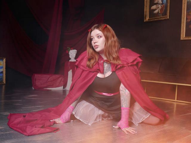 YoureGift, a pale-skinned woman with red hair, kneels on a wooden floor wearing a red cape, black dress, and pink gloves. She has tattoos on her arms, and red devil horns on her head. Her dress has a white lace skirt, and she's looking to the left. The room has dark walls, red drapes, and a white bust on a pedestal in the background. A gold-framed painting is partially visible on the right. The lighting is soft, creating a slightly ethereal effect.