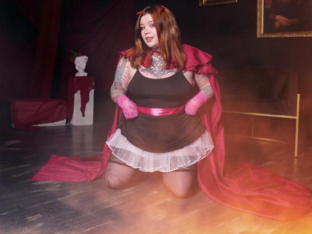 YoureGift, a chubby woman with red hair, wearing a red cape, black dress with white lace, pink gloves, and red belt, kneeling on wooden floor, showing tattoos.