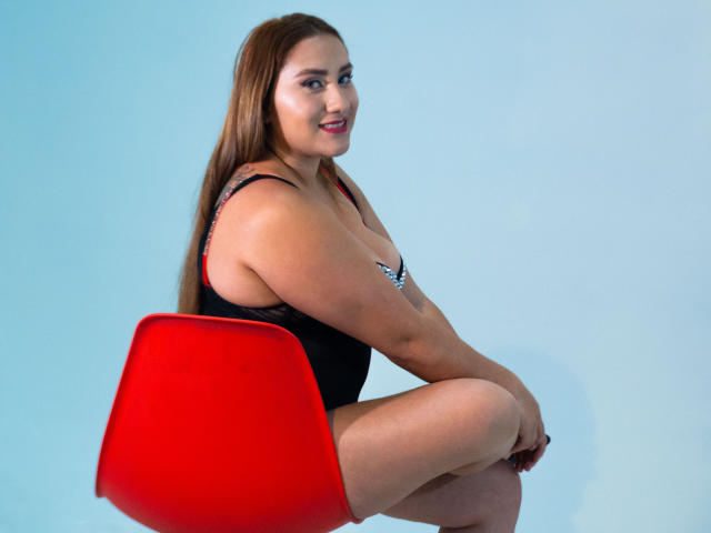 SamanthaWax with long brown hair, black lingerie, and red chair, sitting with legs crossed, showing her ass and tits, smiling at the camera.