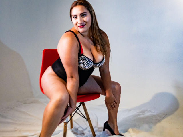 SamanthaWax with big boobs, wearing a black and white studded bra, black thong, and black heels, sitting on a red chair, smiling, showing her ass, in a studio.