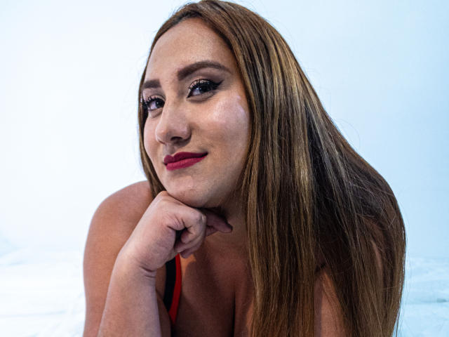SamanthaWax with long brown hair, red lipstick, and black top, resting her chin on her hand, smiling subtly, showing her cute face and small, perky tits.