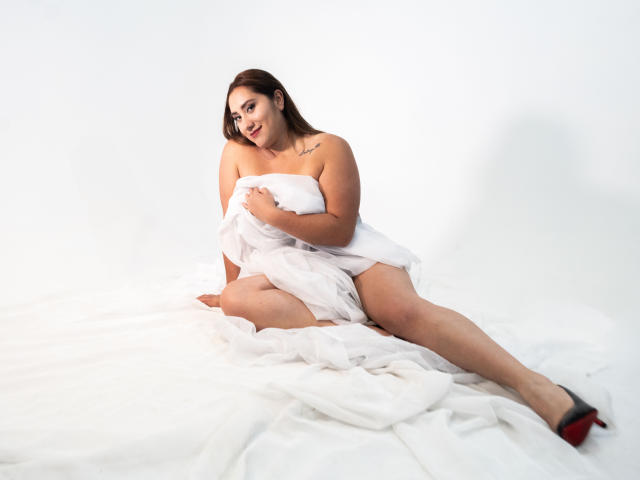 SamanthaWax with large tits, curvy ass, and a tattoo on her right shoulder, sitting on white fabric, holding a towel over her pussy, wearing red high heels.
