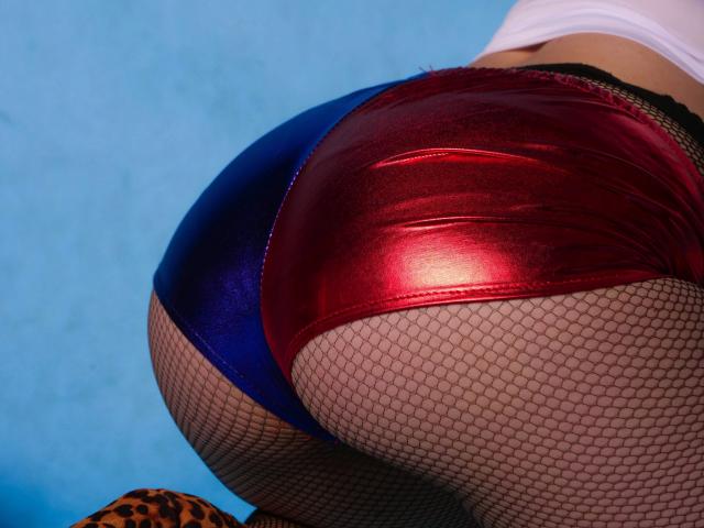 WildeDesire's shiny red and blue metallic butt shorts, tight against her round ass, over fishnet stockings, with a leopard print object at the bottom. Blue background.