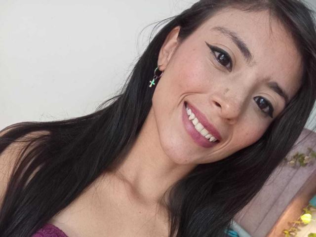 AlanaMoore has long black hair, brown eyes, and is smiling with pink lipstick, wearing green cross earrings, and showing white teeth. Her tits are not visible.