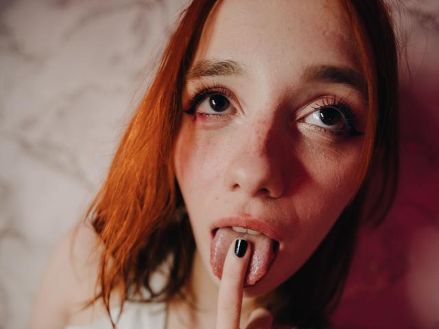 DarkWilllow has red hair, freckles, and is sticking her tongue out with her finger touching it. She has black nail polish and is looking up with big eyes.