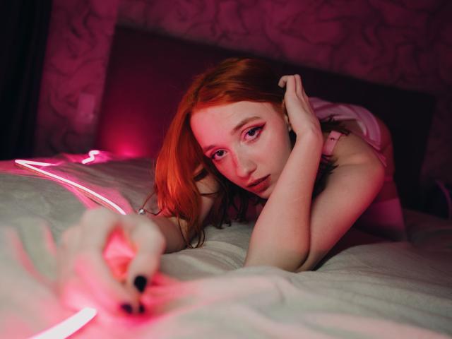 DarkWillow with red hair, pink eyeshadow, and black nails, laying on a bed, looking seductively at a pink neon light, her hand touching her ear.