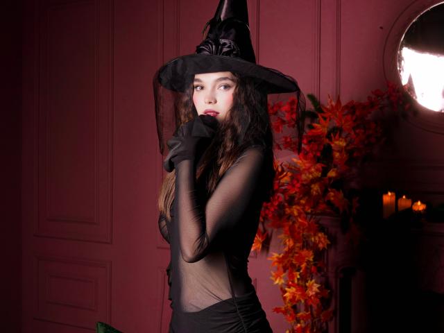 OwnPath stands center-frame, wearing a black witch hat with a ribbon and a sheer black dress that reveals her skin underneath. She has long, wavy brown hair, pale skin, and dark red lipstick. Her right hand, gloved in black, touches her lips. The background has a red wall and a round mirror on the right, with autumn leaves and candles on a dark shelf. The lighting is soft, highlighting her face and the texture of her dress. The image uses dark, moody colors and has a gothic, mysterious vibe.