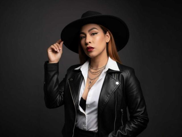SharomCastle with brown hair, wearing a black leather jacket, white shirt, black hat, and necklaces, holding hat, red lipstick, dark background, looking confident.