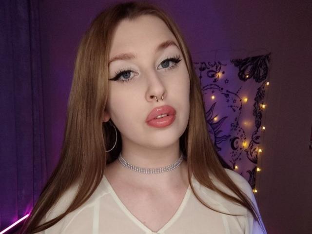 AliceLovels has long brown hair, blue eyes, winged eyeliner, pink lips, nose ring, hoop earrings, and a silver choker. She's wearing a sheer white top.