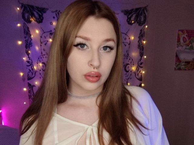 AliceLovels has pale skin, long red hair, blue eyes, and wears a nose ring and choker. She's looking at the camera with full pink lips, wearing a white top.