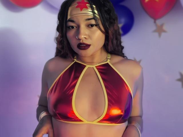 JeanineX with medium tits in a shiny red and yellow Wonder Woman bikini top, dark wavy hair, red star headband, and dark lipstick, standing in a softly lit room.