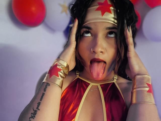 JeanineX with black hair, gold star headband, red and gold costume, tongue out, hands on head, nose ring, tattoo on left arm, looking up with eyes half-open.