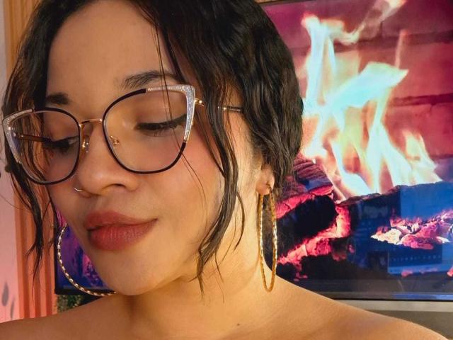 JeanineX with wavy black hair, glasses, nose ring, and gold hoop earrings, smiling with closed eyes, showing her red lipstick, and bare shoulders. TV with colorful flames in background.