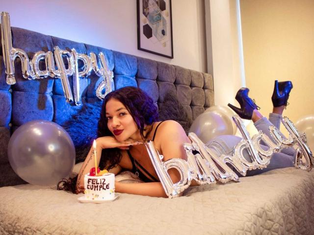 JeanineX with dark curly hair, red lipstick, black lingerie, blue high heels, lying on bed, holding "Felice" cake, silver "Happy Birthday" balloons, gray tufted headboard, white quilt.