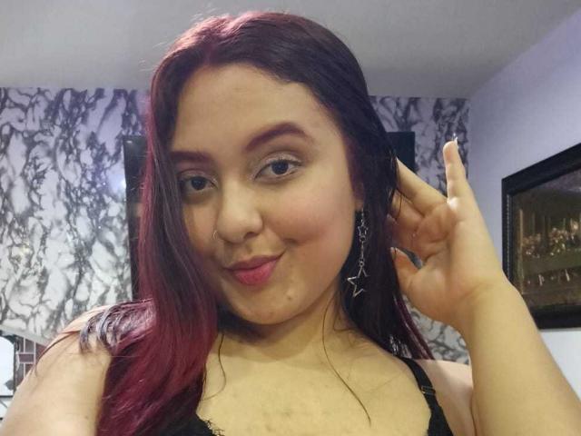 NicolleCollins has red and black hair, a nose piercing, and star earrings, with her hand in her hair, smiling slightly, wearing a black top, showing her tits.