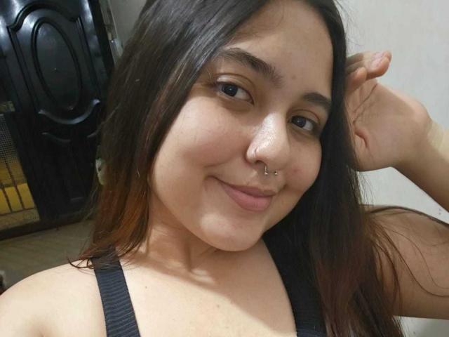 NicolleCollins with brown hair, wearing a black tank top, smiling, nose pierced, right hand in hair, slightly flushed skin, soft brown eyes, and a relaxed, cute expression.