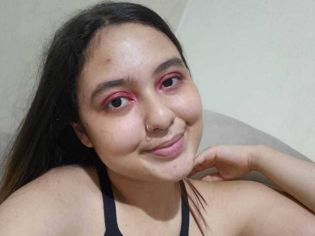 NicolleCollins has pink eyeshadow, a nose ring, and a small pimple on her forehead, smiling with long brown hair, wearing a black tank top, and resting on a couch.