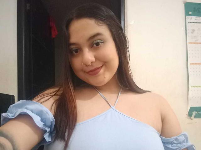 NicolleCollins has light skin, brown eyes, and a nose ring. She's wearing a light blue off-shoulder top, smiling at the camera with her brown hair. Boobs visible.