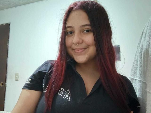 NicolleCollins has red and black dyed hair, a nose ring, and a small smile, wearing a black shirt, showing her tits slightly under the shirt's collar.