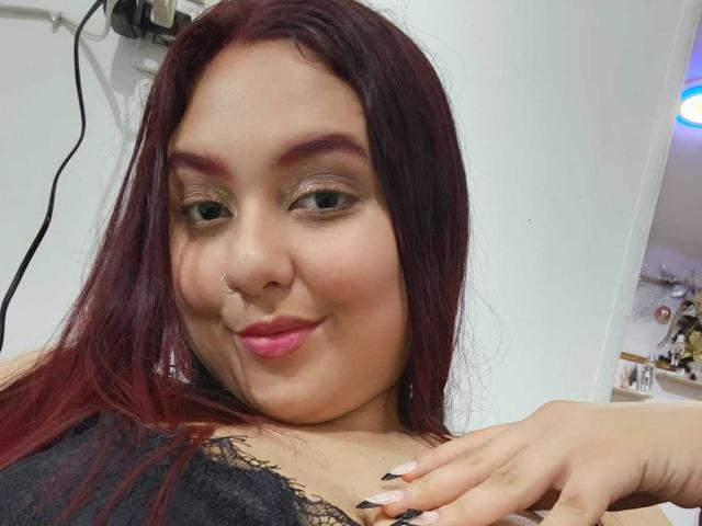 NicolleCollins has dark red hair, a nose ring, pink lipstick, and black lace top, smiling with her hand on her chest. Her nails are long and painted black.