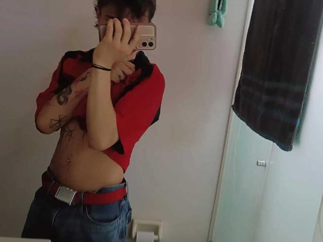 Jeromxx, vestindo uma camisa vermelha e preta, levantando-a para mostrar uma barriga nua, tatuada e com piercings, e usando jeans azuis com um cinto vermelho, tirando uma selfie no espelho..
