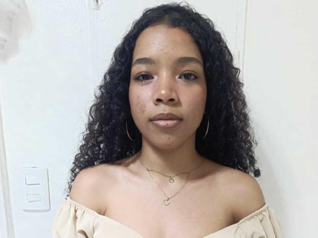 BettyBrunette69 has curly black hair, tan skin, small nose, and is wearing an off-shoulder beige top with gold hoop earrings and a necklace. Her pussy is not visible.