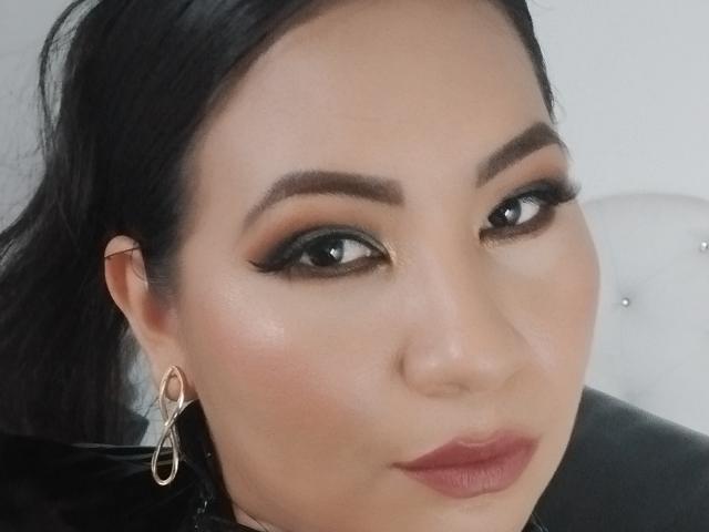 DominikaR has dark, thick eyebrows, black eyeliner, and smoky eye makeup. She's wearing a gold hoop earring on her left ear and has pink lipstick on her pouty lips.