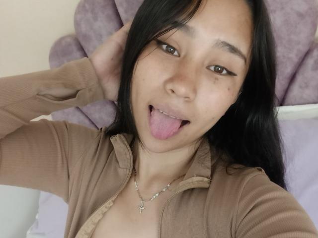 Karollteyes tiene el pelo largo y negro, ojos marrones, y está sacando su lengua rosa. Lleva una camiseta beige con cremallera y un collar de plata con una cruz..