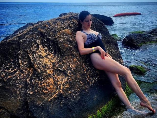 Diamond69s with big ass, small tits, and a purple leopard-print one-piece swimsuit, leaning against a rock by the ocean, legs crossed, wet skin, dark hair, yellow bracelet.