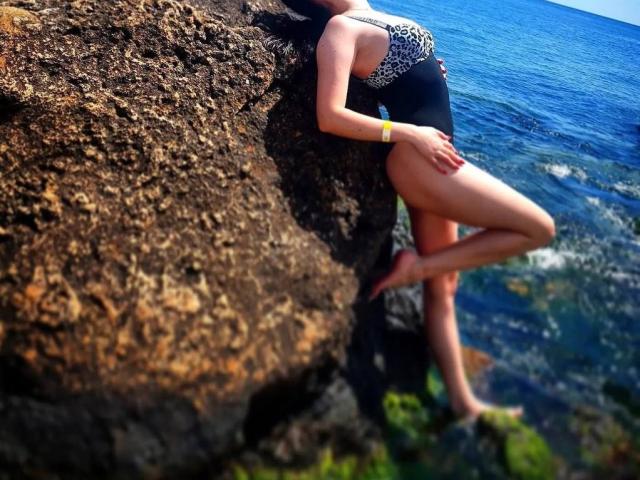 Diamond69s leans against a rock, wearing a black and leopard-print one-piece, showing her ass, with a yellow bracelet on her right wrist, by the blue ocean.