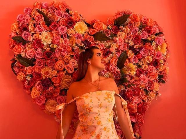 Diamond69s with long brown hair, off-shoulder floral dress, standing in front of massive heart-shaped flower wall, pink, red, and orange roses. Her tits are slightly visible.