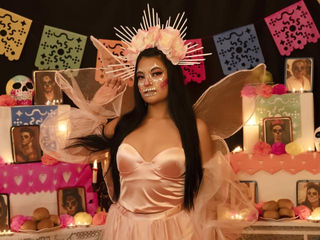 JulietaRizzo with long black hair, wearing a pink satin dress, fairy wings, and a sun crown, has sparkly face paint and large pink-tinted eyes, standing in a festive setup.