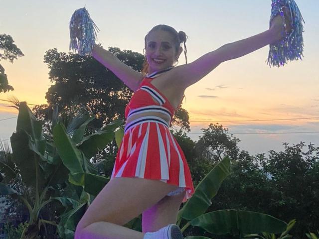EliBaker with arms raised, wearing a red and white cheerleading outfit, holding pom-poms, showing her tits and ass, standing in front of a sunset with trees.