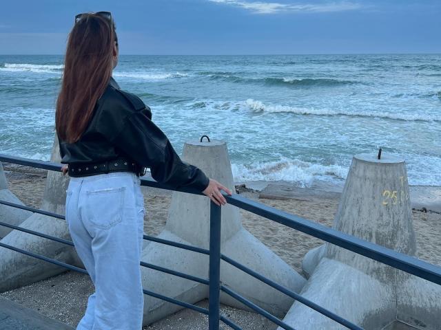 Diamond69s with long brown hair, black leather jacket, and white jeans, stands against a metal railing, looking at the ocean, butt slightly raised, hands on railing.