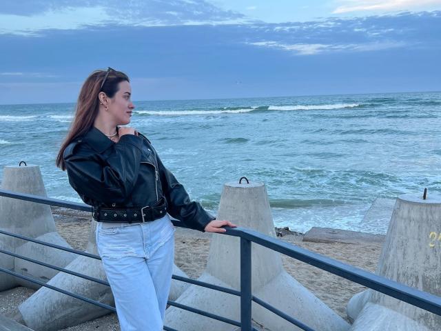 Diamond69s stands on a beach, wearing a black leather jacket, white pants, and a belt, leaning on a concrete railing, looking at the ocean with a relaxed expression.