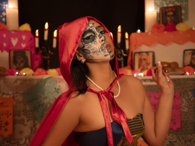 KattyRivera with skull face paint, red hood, pearl necklace, and strapless blue top, holding up finger, showing off tits, in dimly lit room with candles and decorations.