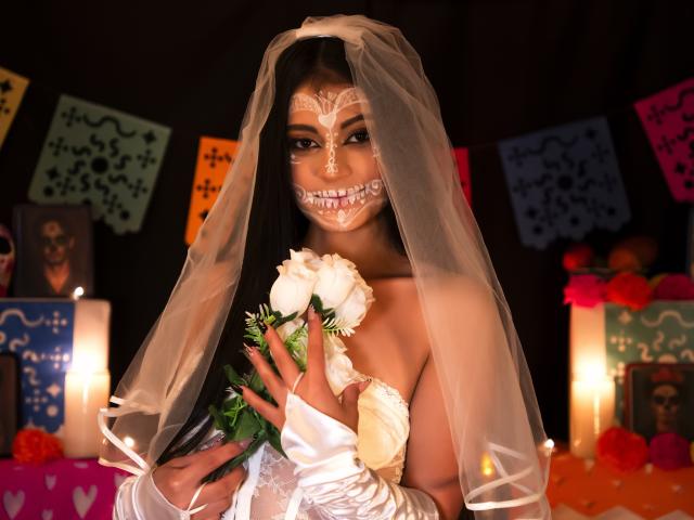 LizzaRose in a white wedding dress with a veil, face painted like a sugar skull, holding white roses, wearing satin gloves, dark hair, smiling, black background.