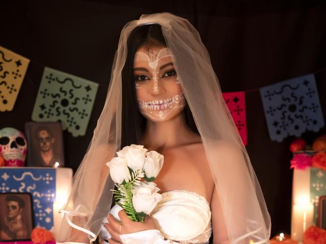 LizzaRose with white face paint, skull design on cheeks, wearing a veil, holding white roses, in a dress, candlelight, dark background, Day of the Dead decor, smiling.