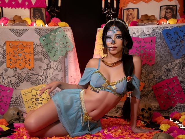 MadissonBlue, con lingerie di pizzo blu, gioielli dorati e trucco sul viso, sdraiata su petali colorati, circondata da teschi e candele. Ha piccoli seni, una vita sottile e lunghi capelli neri..