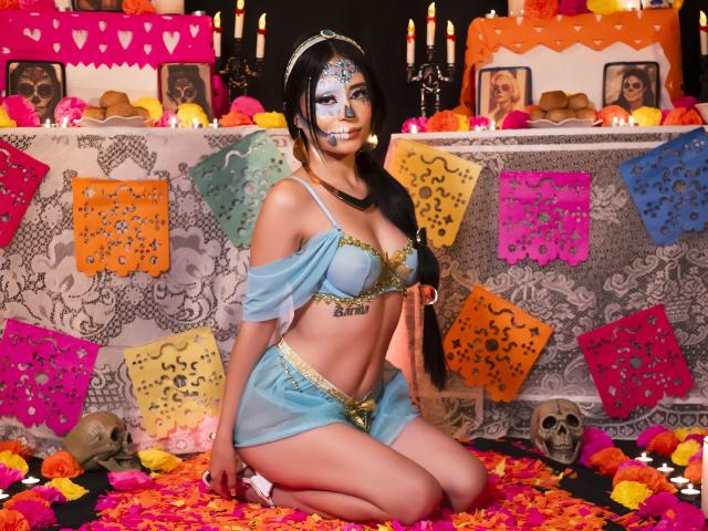 MadissonBlue kneeling, wearing blue and gold lingerie with sheer fabric, face painted in Day of the Dead style, tattoos visible, surrounded by skulls, candles, and colorful paper decorations.