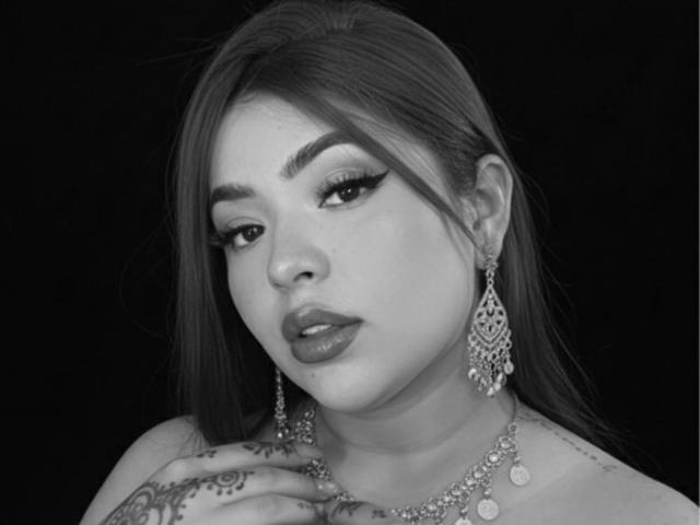 Black-and-white photo of KrishnaHot with long hair, full lips, and dramatic eyeliner. She's wearing ornate earrings, a necklace, and has tattoos on her hand. Boobs partially visible.