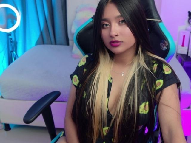 AmmyLewis with long black and blonde hair, wearing a black shirt with green avocado prints, sitting in a gaming chair, pink lipstick, cute, and a small cross necklace.
