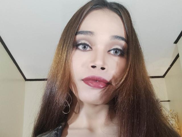 Close-up of DiamondNicky. She has long, straight brown hair with subtle highlights, and gray eyes with heavy black eyeliner. Her full lips are painted a shiny pink-red. She wears a small silver hoop earring on her left side. Her skin is light and smooth. The image focuses on her face, centered in the frame. The ceiling in the background has black trim, contrasting with the beige walls. The lighting is even, highlighting her facial features. Her expression is neutral, slightly parted lips. The overall composition is straightforward, with DiamondNicky as the central subject.