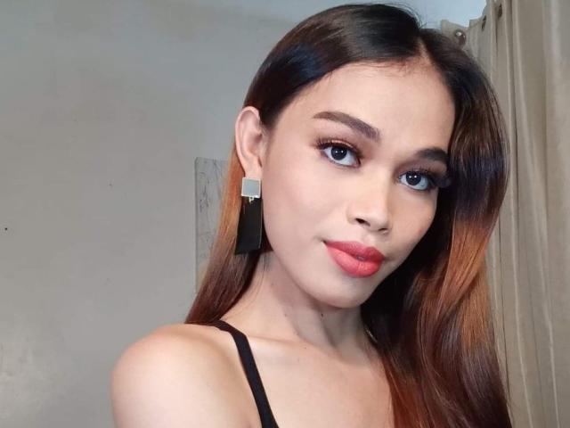 DiamondNicky, a young woman with long brown hair, wears black earrings and a black spaghetti strap top. She has light skin, dark eyebrows, and bright red lipstick. Her blue eyes are slightly upturned. The photo focuses on her upper body and face, with soft, natural lighting highlighting her features. The background is blurred, with beige curtains on the right. The overall composition is simple, with a close-up, slightly angled view.